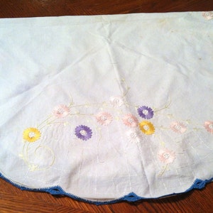 Puede incluir: Un mantel blanco de ganchillo con un borde festoneado azul y flores bordadas en tonos de rosa, amarillo y morado.
