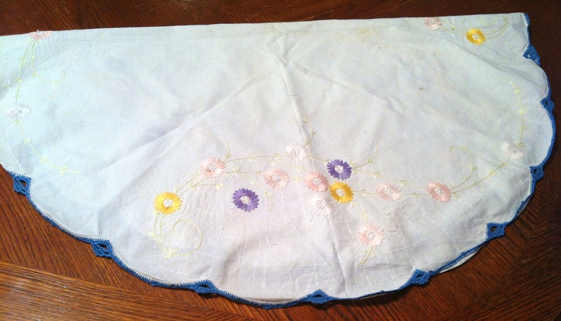 Puede incluir: Un mantel blanco de ganchillo con un borde festoneado azul y flores bordadas en tonos de rosa, amarillo y morado.