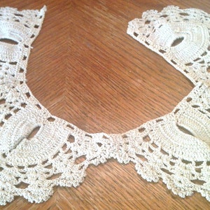 May include: A delicate crocheted lace collar in a cream color. The collar is made of intricate stitches and has a scalloped edge.
