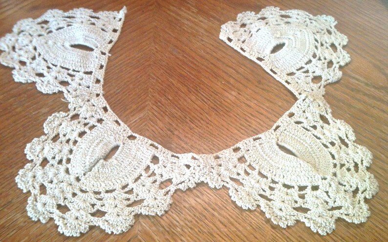 May include: A delicate crocheted lace collar in a cream color. The collar is made of intricate stitches and has a scalloped edge.