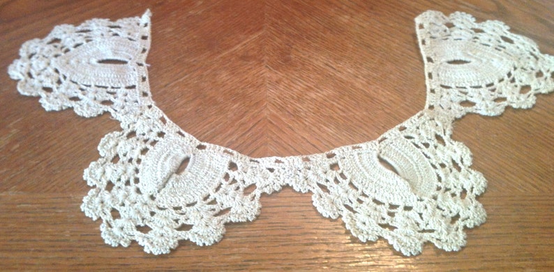 May include: A white crocheted collar with a scalloped edge. The collar is made of delicate lace and has a vintage look.