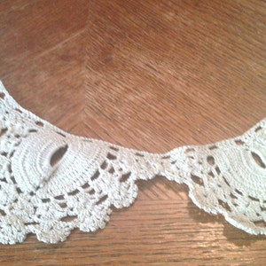 May include: A white crocheted lace collar with a scalloped edge. The collar is made of delicate, intricate stitches and has a vintage look.