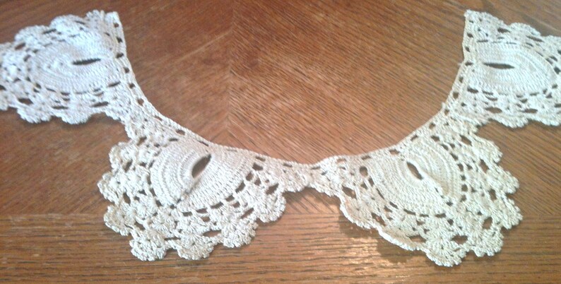 May include: A white crocheted lace collar with a scalloped edge. The collar is made of delicate, intricate stitches and has a vintage look.