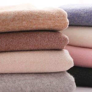 May include: A stack of folded fabric pieces in various solid colors including light orange, medium brown, light beige, gray, blue, white, pink, and black. Each piece of fabric has a soft, fuzzy, and textured surface appearance.