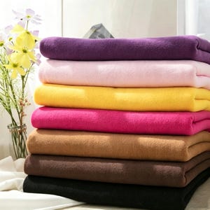 Può includere: Una pila di sette coperte in pile piegate in una varietà di colori solidi tra cui viola, rosa chiaro, giallo, rosa acceso, marrone chiaro, marrone scuro e nero. Le coperte sono impilate ordinatamente, mostrando un tessuto morbido e strutturato.