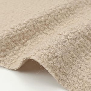 Puede incluir: Una tela de color beige con una superficie densa, rizada y texturizada. El material presenta una construcción de pelo elevado y consistente que crea una apariencia suave y táctil en todo el tejido plegado, con un tono tostado claro.