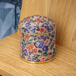 May include: A cylindrical metal canister with a removable lid, featuring a beige background decorated with a dense pattern of small red, pink, purple, and blue flowers with green leaves, finished with a thin gold-toned metal rim at the base.