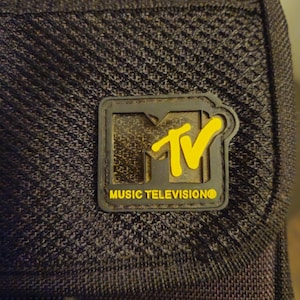 May include: A black textured fabric bag featuring a rectangular rubber patch with the MTV logo in yellow and black. The logo includes the stylized letters MTV and the text MUSIC TELEVISION in yellow capital letters against a dark background.