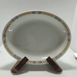 Może przedstawiać: Owalny biały półmisek ceramiczny z ozdobnym wzorem na obrzeżu, składającym się z naprzemiennych złotych kłosów zbóż oraz małych czarnych i złotych geometrycznych kwadratowych motywów, ułożonych w powtarzający się wzór wokół całego brzegu.