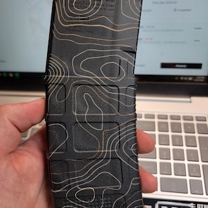 Puede incluir: Un cargador de rifle de polímero negro con un patrón de líneas topográficas de color canela. La superficie tiene texto en relieve que incluye 5.56x45, Magpul Industries Corp, Cage 1LX50, Made in USA e IP.MAGPUL.COM. Posee un agarre texturizado.
