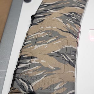 May include: A polymer rifle magazine featuring a tiger stripe camouflage pattern in shades of tan, brown, grey, and black. The rectangular magazine has a textured surface with a grid pattern at the base and a slightly curved profile for ammunition.
