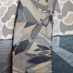 May include: A polymer rifle magazine featuring a geometric camouflage pattern in shades of tan, gray, and black. The surface displays molded text including 5.56x45, GEN M2, and PMAG 30, with a textured grip area and a rectangular base plate design.
