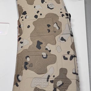 Puede incluir: Un cargador de rifle con un patrón de camuflaje en beige, marrón y negro. El cargador tiene una superficie texturizada con secciones de rejilla rectangulares y un pequeño logotipo circular cerca de la parte superior. El diseño cubre todo el cuerpo visible.