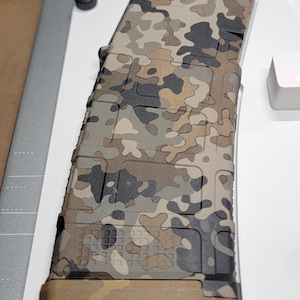 Pode incluir: Um carregador de rifle com um padrão de camuflagem multicolorido em tons de bronzeado, verde-oliva, cinza-escuro e preto. O carregador tem uma superfície texturizada com um padrão de grade e uma placa de base sólida na cor bronzeada na base.