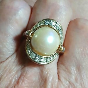 May include: A gold-toned ring featuring a large, round, cream-colored pearl at the center. The pearl is surrounded by an oval-shaped frame embedded with small, clear, sparkling stones that catch the light, creating a bright and reflective appearance.
