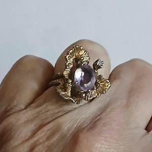 May include: A gold ring featuring a central oval-cut light purple gemstone. The setting has a textured, leaf-like design surrounding the stone, with a small, clear, round-cut accent stone positioned on the upper right side of the main setting.