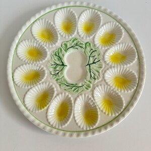 Puede incluir: Una bandeja de cerámica redonda para servir huevos con doce hendiduras ovaladas con centros amarillos dispuestas en círculo. El centro del plato muestra un diseño de hojas pintadas en verde que rodea una zona lisa con borde festoneado.