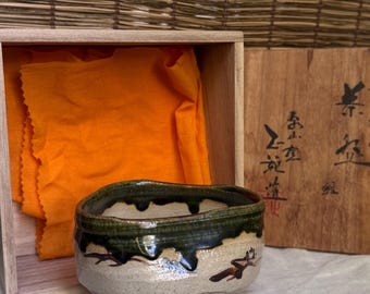 May include: A ceramic tea bowl with a speckled beige base and a dark green drip glaze along the rim. The bowl features small brown painted branch motifs and sits in a wooden box lined with a bright orange cloth, with Japanese calligraphy on the lid.
