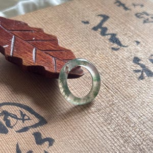 Peut inclure: Une bague circulaire translucide présentant des inclusions vertes semblables à de la mousse dans tout le matériau. L'anneau possède une surface lisse et polie ainsi qu'un profil arrondi, révélant des variations naturelles de couleur.