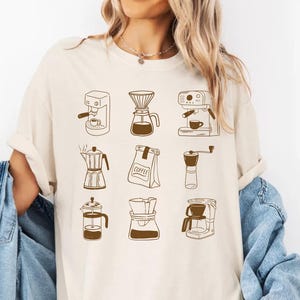 May include: An ivory t-shirt featuring a brown graphic print of nine different coffee brewing methods and equipment, including a coffee bag labeled COFFEE, arranged in a grid pattern. The shirt has short sleeves and a crew neckline.