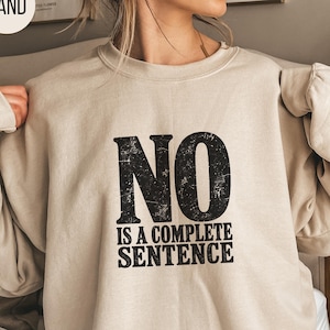 May include: A sand-colored crewneck sweatshirt featuring the black distressed-style text NO IS A COMPLETE SENTENCE printed on the front. The garment has long sleeves and a ribbed neckline, providing a simple and straightforward design.