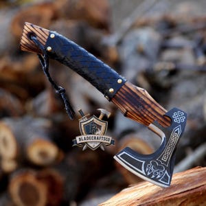 May include: A hand axe with a dark, scorched wooden handle wrapped in a black braided leather grip secured by brass rivets. The metal axe head features an engraved bear paw print and intricate knotwork patterns, with a leather lanyard attached.