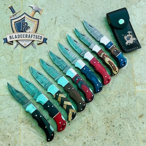 May include: A collection of eight folding pocket knives with Damascus steel blades and multi-coloured wooden handles, arranged diagonally. A black leather sheath with a silver snap button and the BLADECRAFTSCO logo is positioned at the top right.