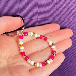 May include: A circular keychain featuring a repeating pattern of small, round beads in alternating colors of hot pink, metallic gold, white, and beige. The beads are strung on a flexible cord and attached to a silver-toned metal split ring.