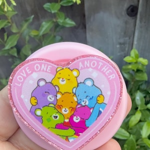 May include: A circular pink phone grip featuring a heart-shaped topper with a glittery border. The center displays a group of five colorful cartoon bears hugging, with the text LOVE ONE ANOTHER printed in white along the top edge of the heart.