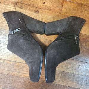 May include: A pair of dark brown suede ankle boots featuring a block heel, a rounded toe, and a silver-toned metal zipper closure on the outer side of each boot. The boots have a clean, solid-colored exterior with visible stitching along the seams.