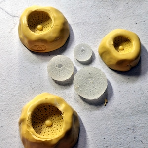 May include: Three yellow silicone moulds with a textured, porous interior surface and a small central protrusion, accompanied by three translucent, circular resin discs featuring a fine, dotted pattern on their flat surfaces.