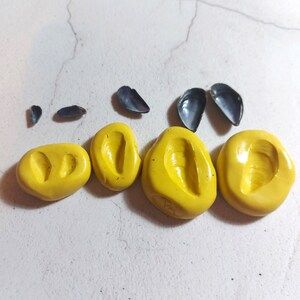 May include: Five yellow silicone moulds for making resin jewellery. The moulds are shaped like seashells and are arranged in a row on a white surface.