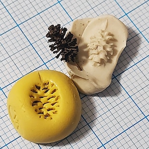 May include: Two clay impressions of a pine cone, one yellow and one white. The yellow impression is raised and the white impression is recessed.