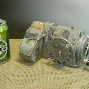 Puede incluir: Una calculadora mecánica vintage de color plateado con manivela, dial numérico circular y botones numerados, colocada junto a una lata de cerveza Tuborg verde de 330 ml para escala. El dispositivo presenta un acabado metálico y manual.