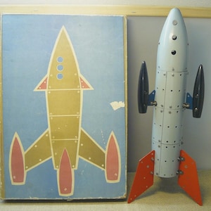 Peut inclure: Une fusée jouet en métal bleu clair avec des ailerons latéraux noirs et des ailerons de base orange, à côté d'une boîte en carton rectangulaire illustrée d'une fusée dorée et rouge sur fond bleu, avec des rivets et des hublots visibles.