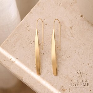 Puede incluir: Un par de pendientes colgantes de tono dorado con un diseño de barra de metal larga, delgada y curva con un acabado liso. Los pendientes están suspendidos de un gancho de alambre fino y arqueado. El texto STELLA BOHEME FINE JEWELRY es visible.