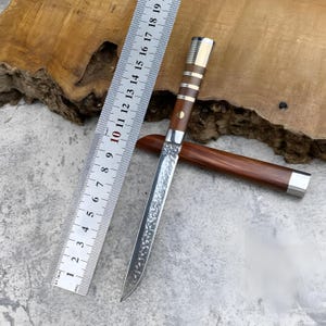 May include: A fixed-blade knife with a textured, hammered metal finish on the blade, a brown wooden handle with silver-coloured metal accents, and a matching wooden sheath. A metal ruler shows the knife length is approximately 19 cm.