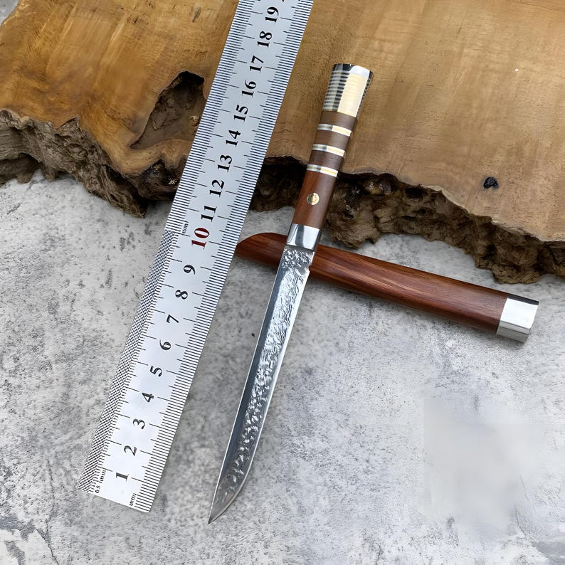 May include: A fixed-blade knife with a textured, hammered metal finish on the blade, a brown wooden handle with silver-coloured metal accents, and a matching wooden sheath. A metal ruler shows the knife length is approximately 19 cm.