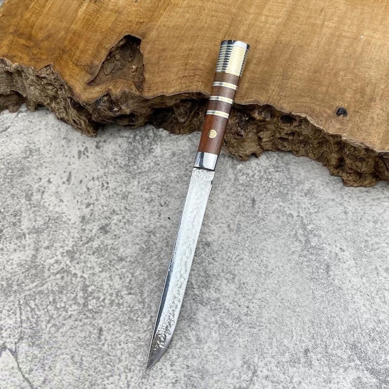 May include: A slender knife with a patterned Damascus steel blade and a cylindrical handle made of dark brown wood, featuring three horizontal silver-coloured metal bands and a small circular inlay near the bolster of the handle.
