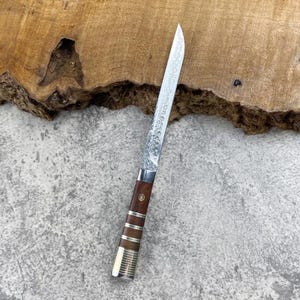 May include: A straight-edged knife with a long, slender, textured metal blade. The handle is crafted from dark brown wood, accented with three horizontal silver-coloured metal bands and a small circular inlay near the bolster area.