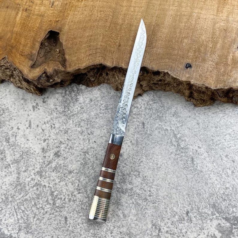 May include: A straight-edged knife with a long, slender, textured metal blade. The handle is crafted from dark brown wood, accented with three horizontal silver-coloured metal bands and a small circular inlay near the bolster area.