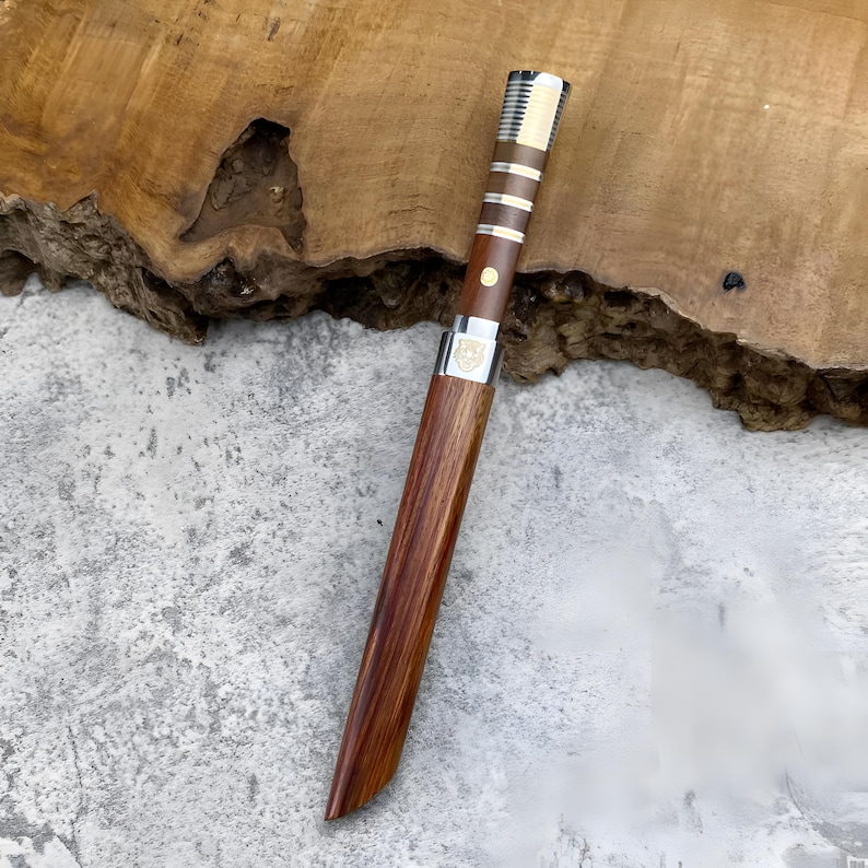 May include: A straight knife with a wooden handle and a matching wooden sheath. The handle features alternating dark wood and metallic silver bands, a single brass pin, and a silver bolster with a small engraved emblem near the blade base.
