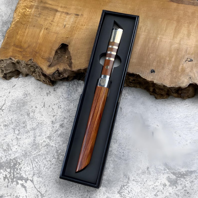 May include: A straight-edged knife with a polished wooden handle and a matching wooden sheath, presented in a black rectangular display box. The handle features silver-coloured metal bands and a small circular emblem near the bolster.