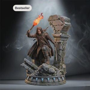Peut inclure: Une figurine détaillée d'un personnage aux cheveux longs portant un manteau marron et tenant une torche enflammée, posée sur un socle en pierre avec des runes gravées. Le socle présente une colonne brisée avec des lianes. Le texte Bestseller est visible.