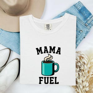 May include: A white short-sleeve t-shirt featuring the black text MAMA FUEL and a graphic of a teal coffee mug with steam rising from it. The shirt has a Comfort Colors brand label at the inner neckline and is displayed with a hat and jeans.