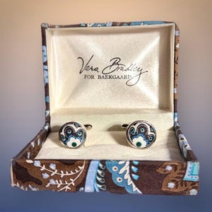 May include: A pair of round cufflinks featuring a brown, blue, and white paisley pattern, presented in a matching fabric-covered box with the text Vera Bradley FOR BAEKGAARD printed in black on the interior cream-colored satin lining.