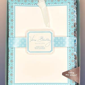 May include: A boxed set of Vera Bradley invitations featuring 12 cards to imprint, quilt embossed sleeves, ribbons, and envelopes. The packaging includes a light blue patterned band with the text Vera Bradley Invitations and a brown corner tab.