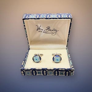 May include: A pair of round silver-tone cufflinks featuring a blue, light blue, and white geometric pattern, presented in a matching patterned box with the text Vera Bradley FOR BAEKGAARD printed on the interior lid lining in black script.