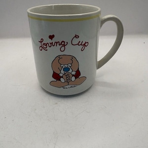 May include: A white ceramic mug featuring a yellow rim and the red text Loving Cup with a small red heart. Below the text is a cartoon illustration of a tan character holding a small white creature with a blue nose, signed by Tom Wilson.