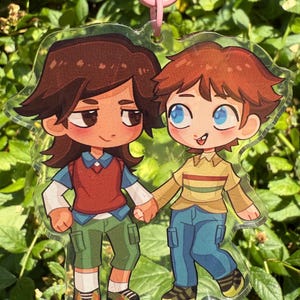 May include: A clear acrylic keychain featuring a cartoon illustration of two characters holding hands. The left character has dark brown hair, a red vest, and green cargo shorts. The right character has light brown hair, a yellow shirt, and blue jeans.